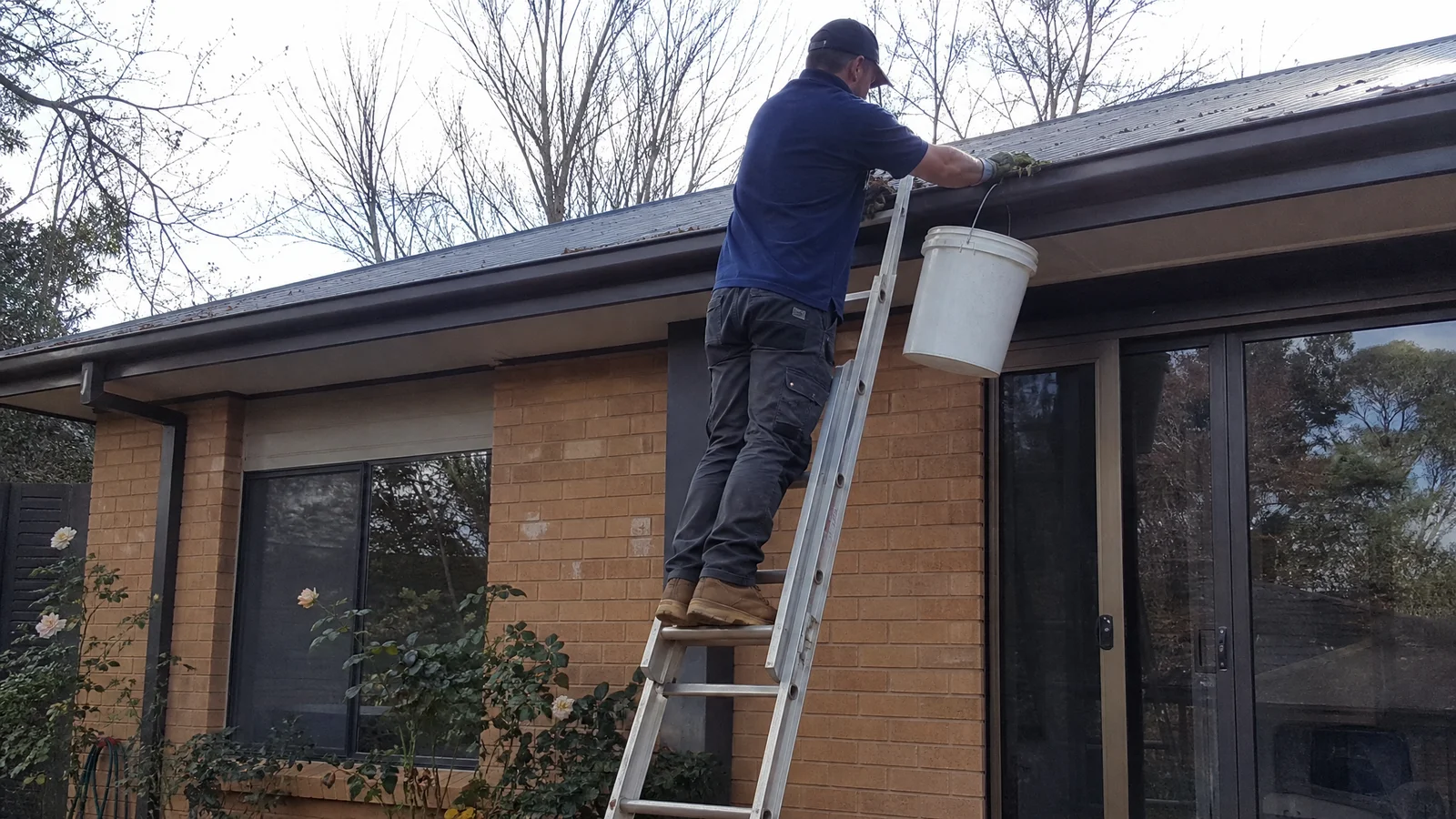Handyman clearing wet leaves from a Colorbond gutter on a Canberra home
